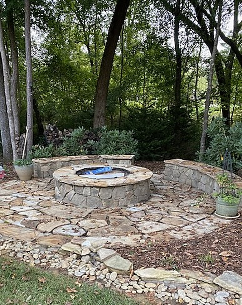 Round stone fire pit with two curved stone seating walls on a flagstone patio ringed with river rock, in a shaded wooded backyard