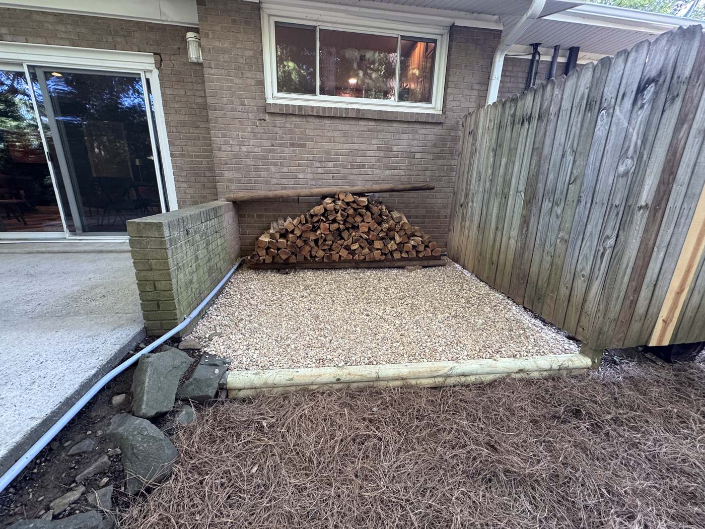 The same corner after installation, with a clean pea gravel pad framed in new timber edging and the firewood restacked