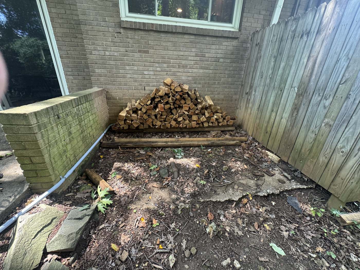 Bare corner between the patio wall and fence with packed dirt, leaves, and old landscape timbers around a firewood pile
