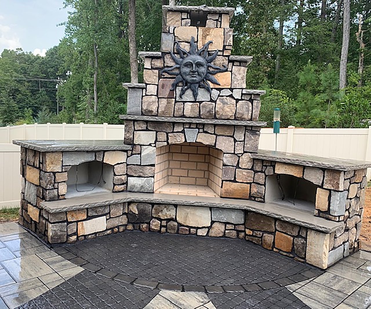 Stone outdoor fireplace with a decorative sun medallion on the chimney and raised hearth benches on both sides, on a patterned paver patio