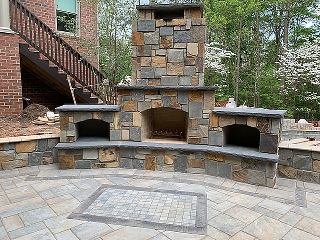 Custom stone outdoor fireplace with firewood boxes on both sides, on a paver patio with a tile mosaic inlay below a deck stair