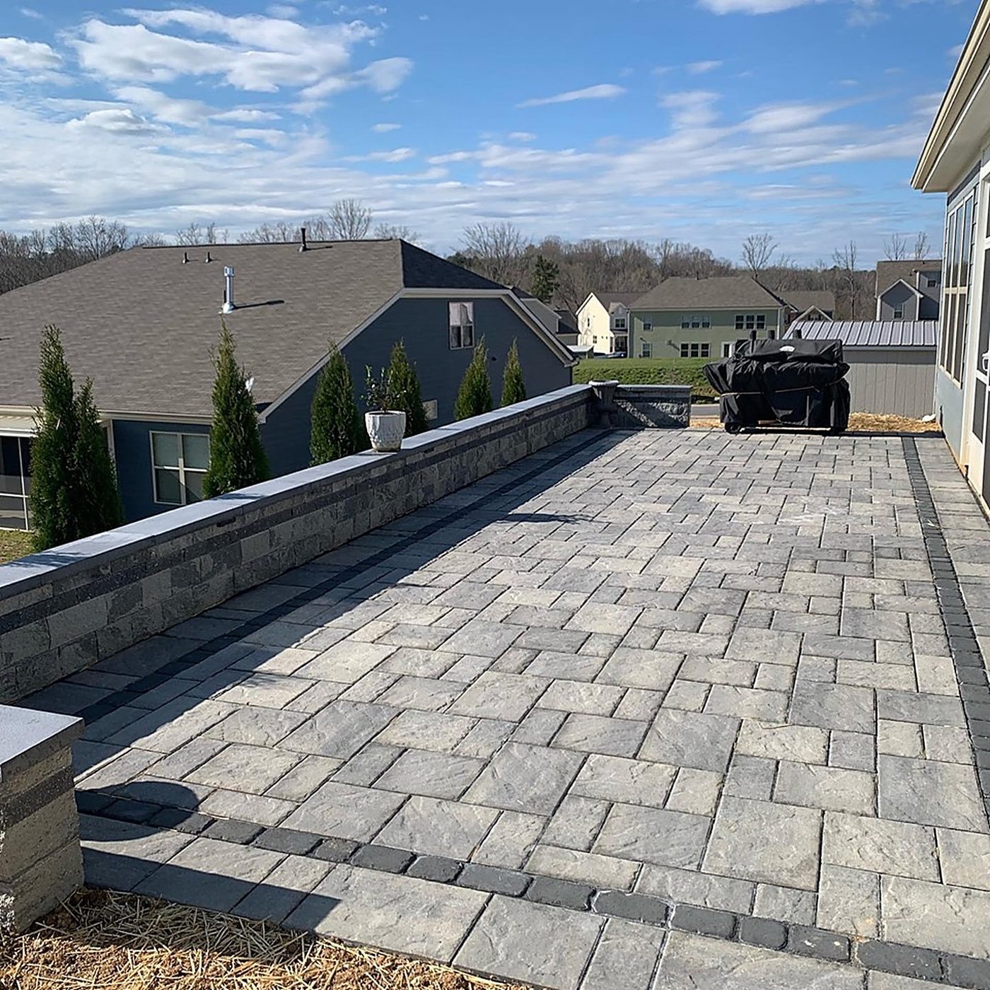 Gray slab paver patio with dark border courses and a block seating wall with stone caps, screened by a row of young arborvitaes