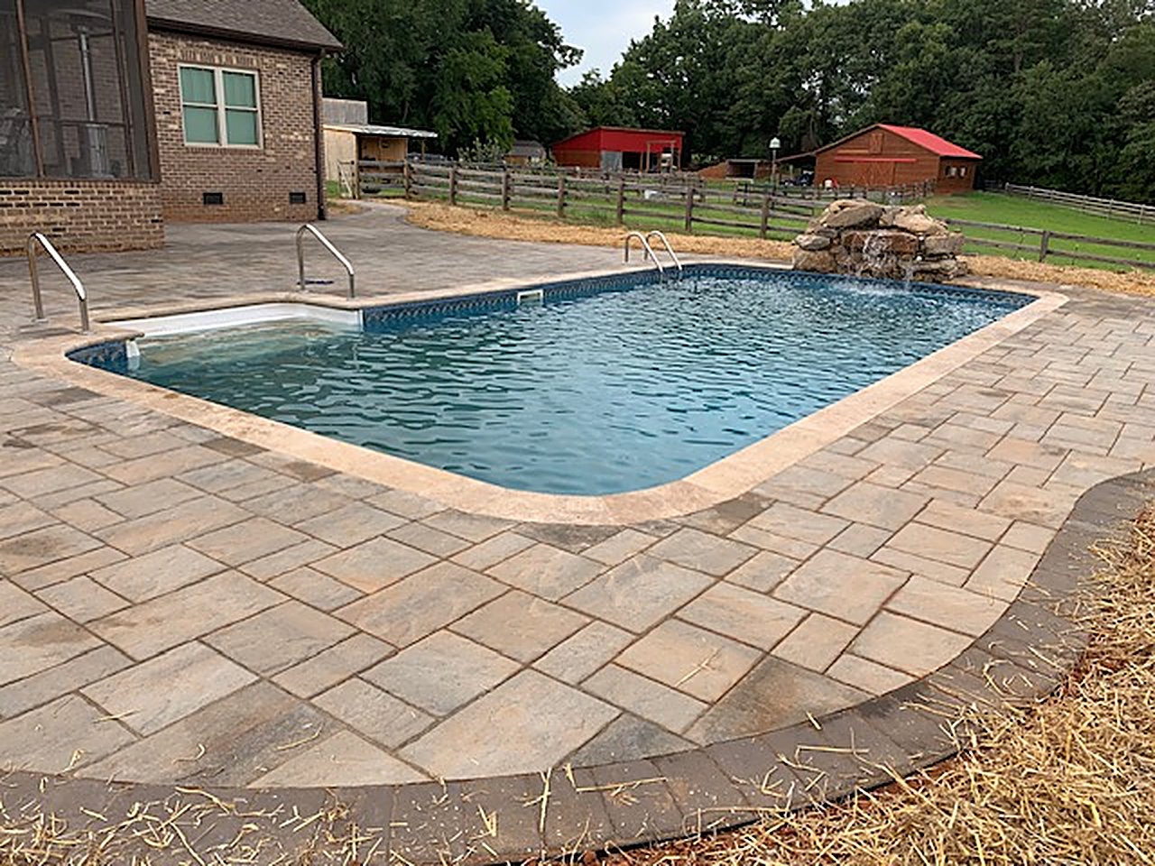 Paver pool deck with light stone coping and a dark border around an in-ground pool with a boulder waterfall feature