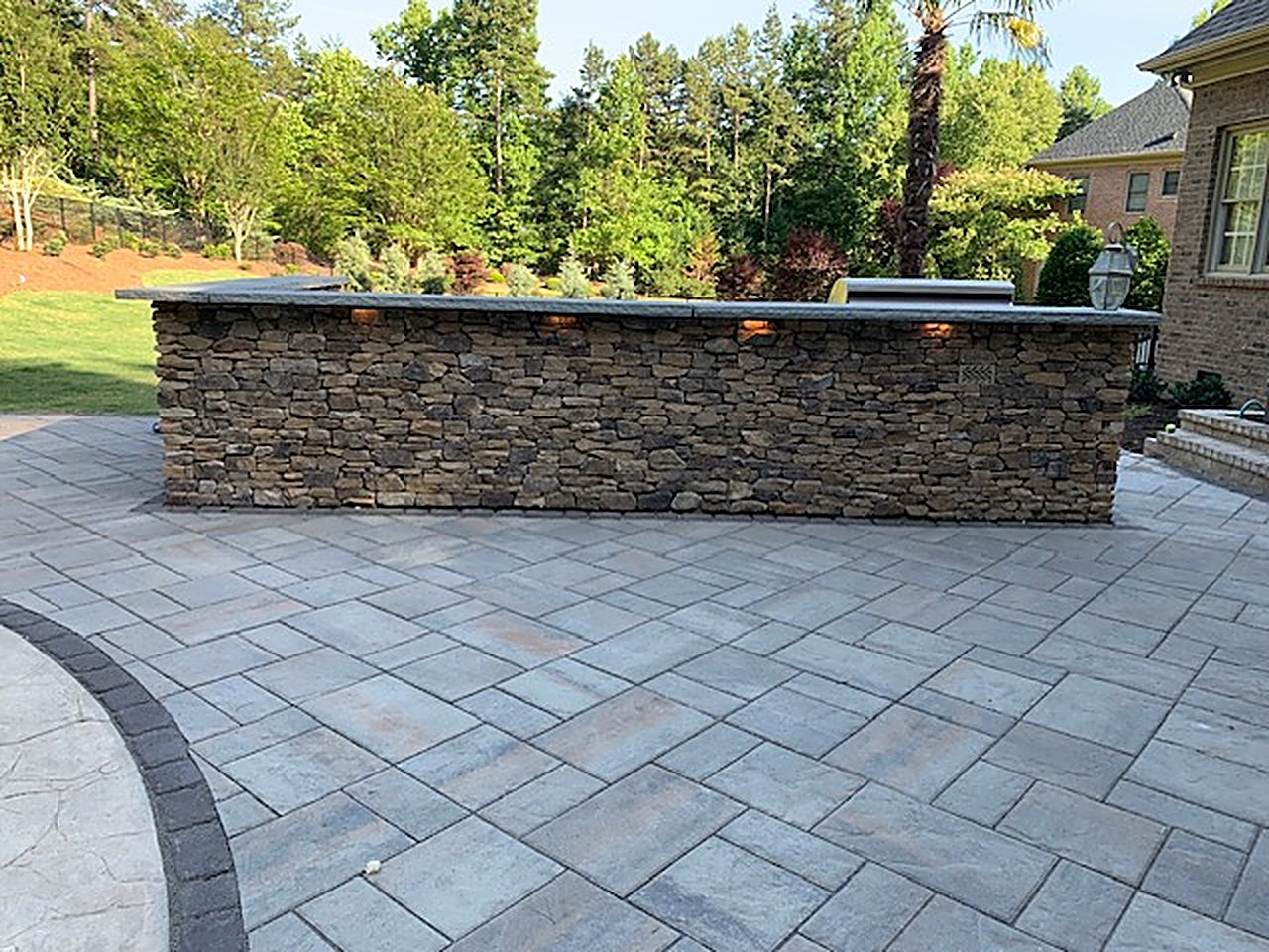 Long stacked-stone outdoor bar with a stone countertop and under-counter lighting on a paver patio at dusk